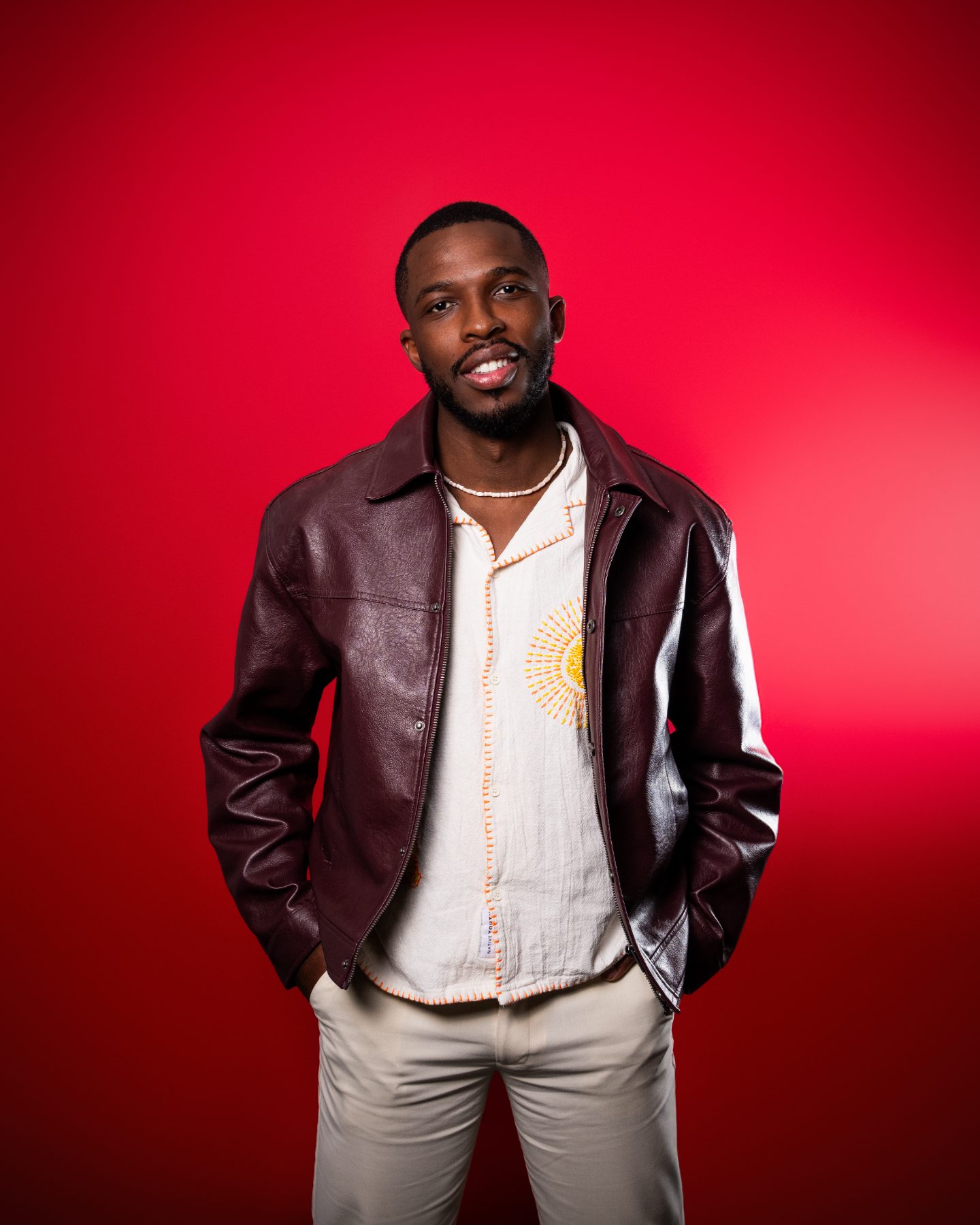 Washington Ali smiling in a burgundy leather jacket on a red backdrop